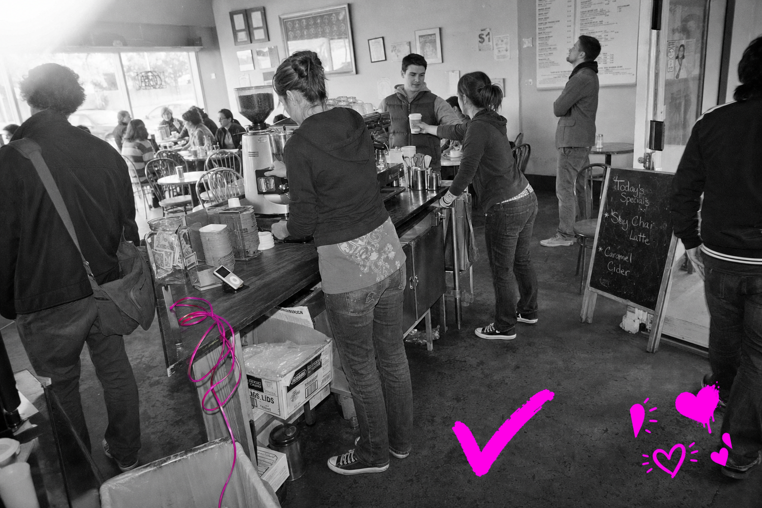 Wide black-and-white point-and-shoot photograph of a busy independent coffee shop interior in 2009, showing staff and customers at a laminate counter and scattered tables, with a coiled power cable near the espresso machine marked in a translucent hot-magenta wash, the only color in the frame.