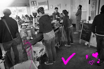 Wide black-and-white point-and-shoot photograph of a busy independent coffee shop interior in 2009, showing staff and customers at a laminate counter and scattered tables, with a coiled power cable near the espresso machine marked in a translucent hot-magenta wash, the only color in the frame.