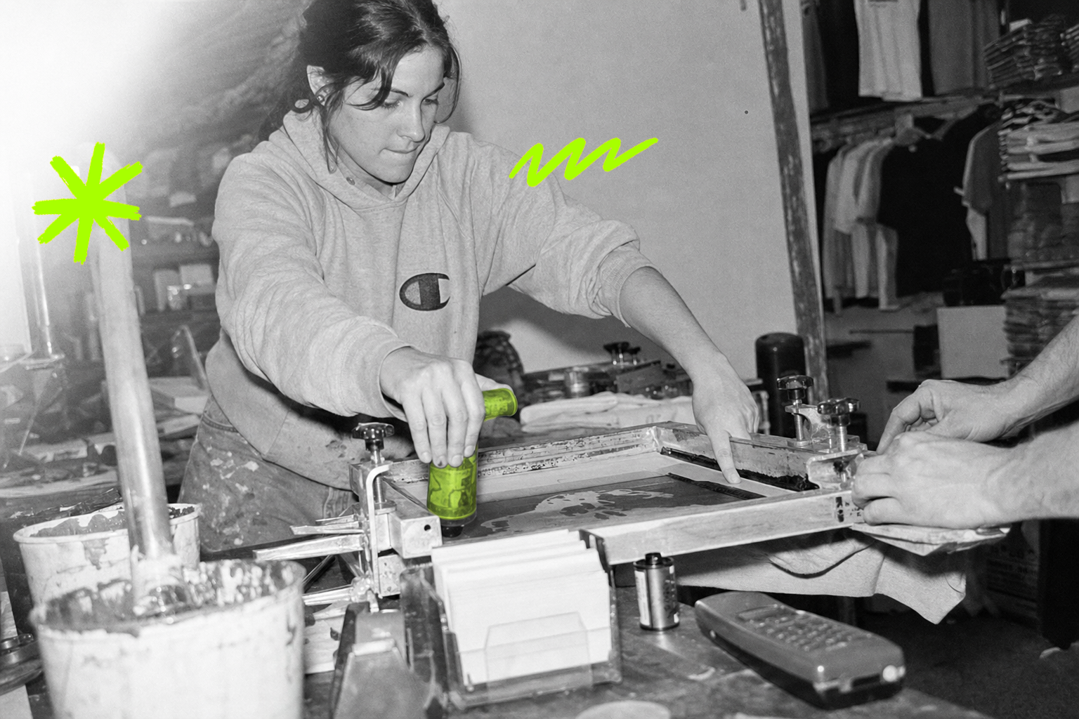 Black-and-white point-and-shoot photograph from 1995 of a young woman pulling a squeegee across a silk-screen frame in a cluttered custom t-shirt shop; the squeegee handle glows with a translucent acid green wash while everything else stays monochrome.