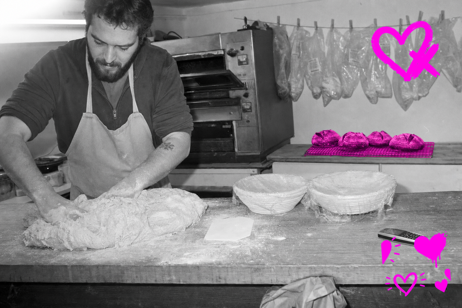 Black-and-white point-and-shoot photograph of a bearded baker in a canvas apron over a hoodie kneading sourdough on a flour-dusted bench in a small bakery, 2007, with a cooling rack of four round loaves glowing in translucent hot magenta behind him.