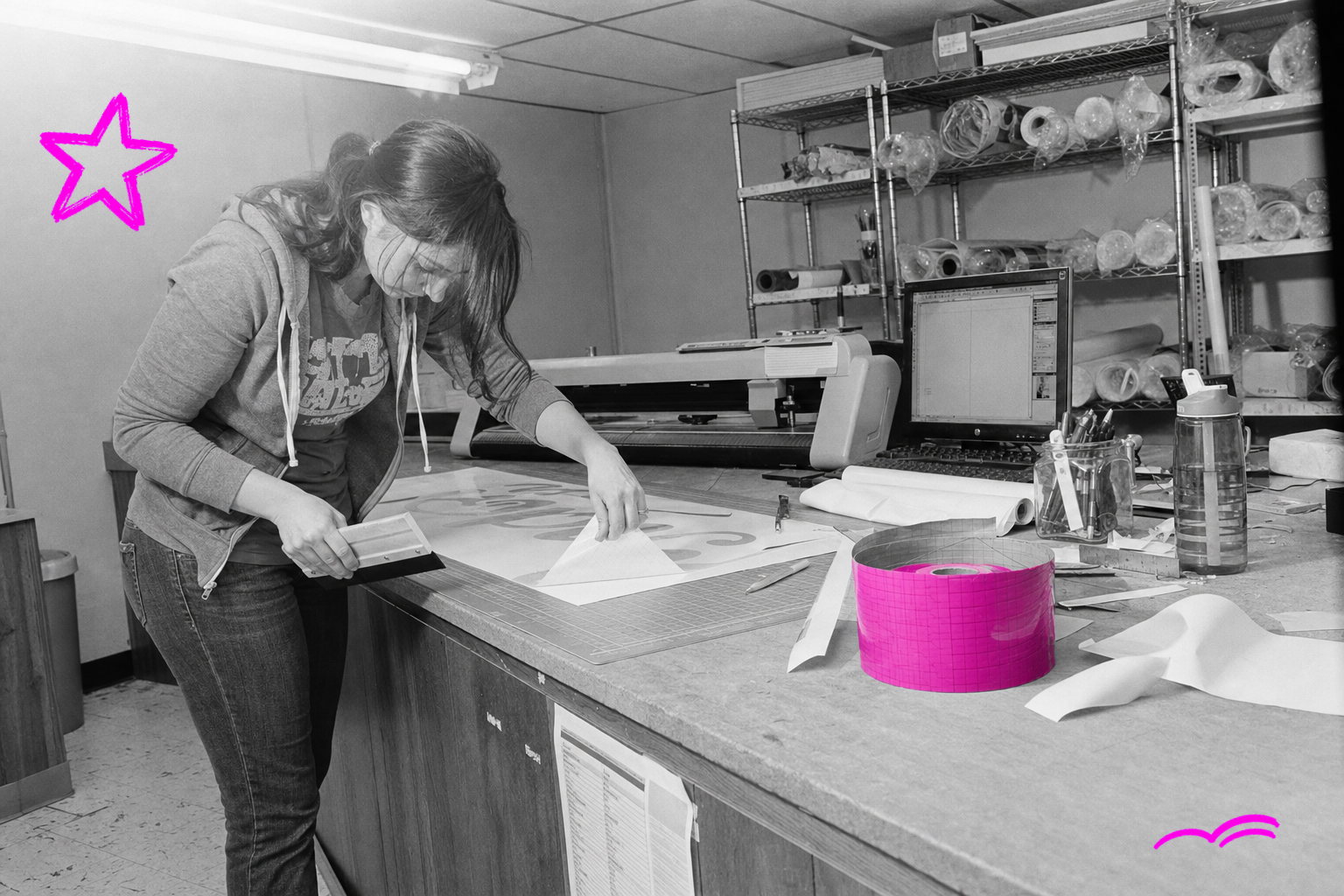Black-and-white snapshot of a young woman in a zip-up hoodie peeling transfer tape off a vinyl lettering panel at a cluttered sign-shop workbench in 2011, a coiled roll of transfer tape in the foreground tinted hot magenta