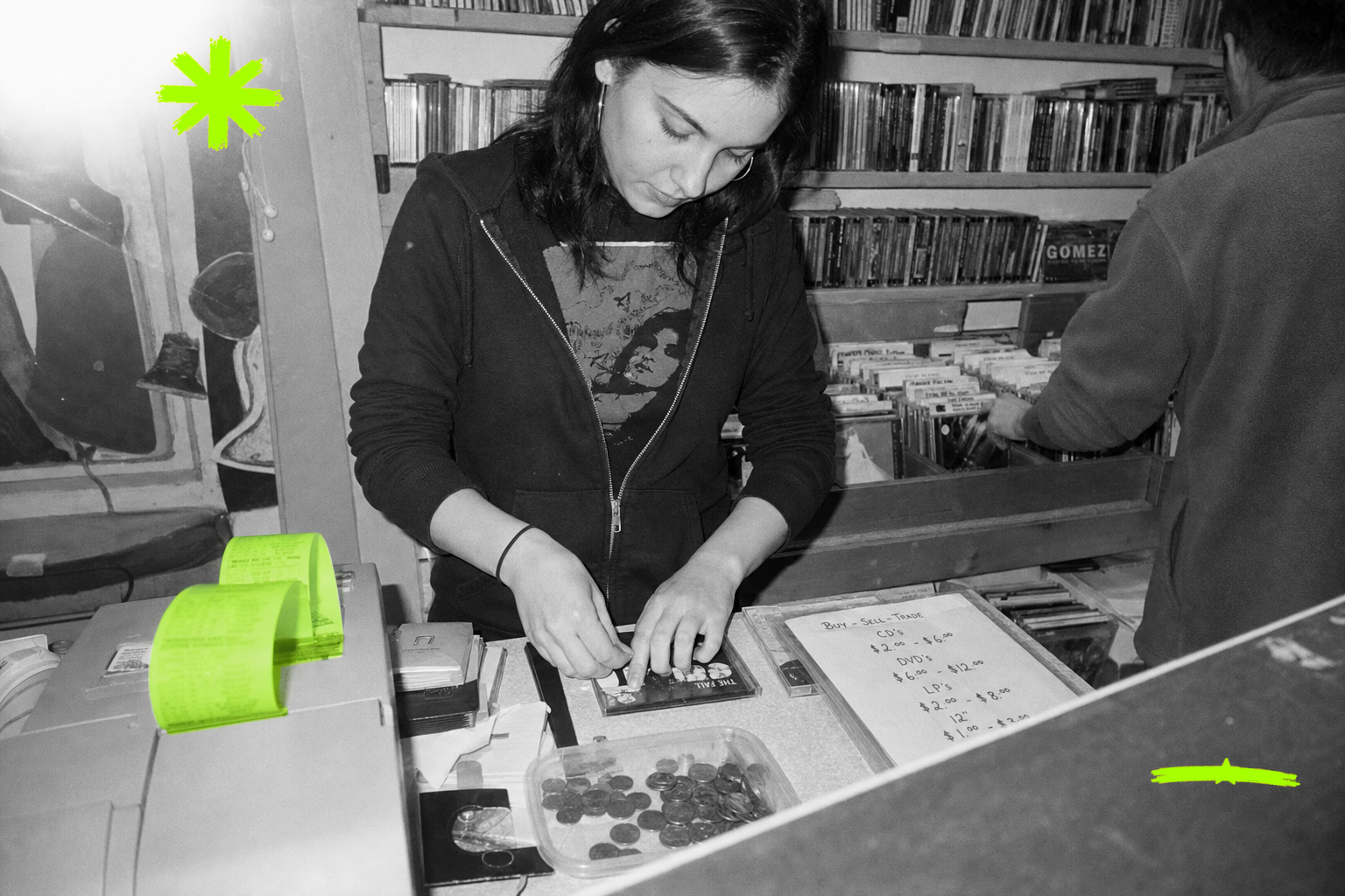 Black-and-white point-and-shoot photo of a young woman behind the counter of an independent record store in 2004, pressing a price sticker onto a used CD sleeve, with a coiled receipt-printer paper roll tinted in acid green on the counter in front of her.