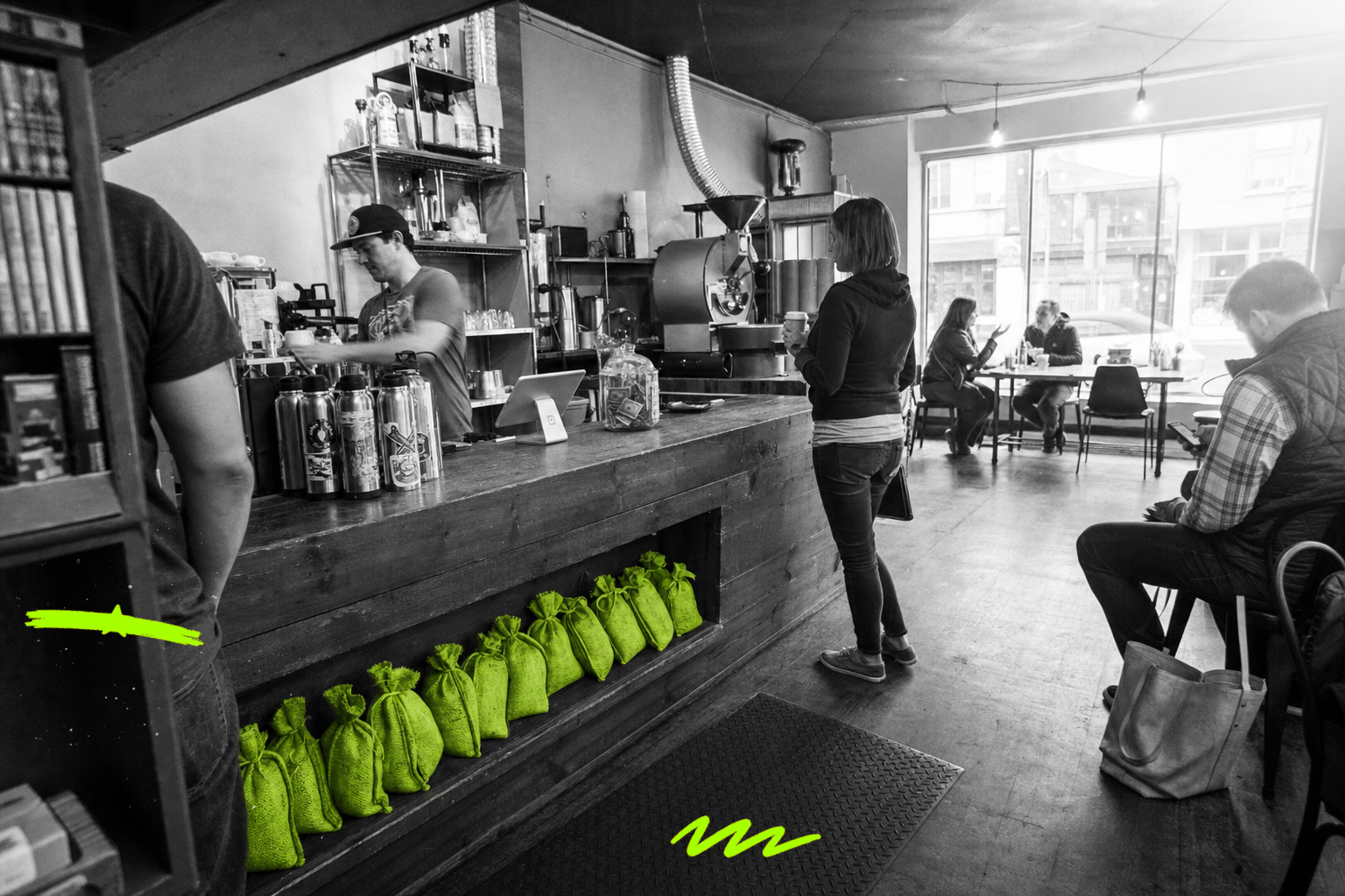 Wide black-and-white point-and-shoot photo inside a busy independent coffee roastery and café in 2013: a barista pulls espresso behind a long counter while customers wait, chat, and scroll phones; a row of small burlap coffee-sample bags on the lower shelf glows with a translucent acid-green wash, the only color in the frame.