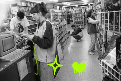 Wide black-and-white point-and-shoot photograph inside a 1997 neighborhood video rental store. A young woman in a zip fleece scans returned VHS tapes at a beige CRT terminal in the foreground while a coworker sorts clamshell cases behind her; two customers browse shelves at mid-distance and a fifth figure disappears toward the back. The coiled barcode-scanner cord glows with a translucent acid-green (#A8FF00) highlighter wash, its ridged texture visible through the color. Direct flash, open shadows, fine film grain.